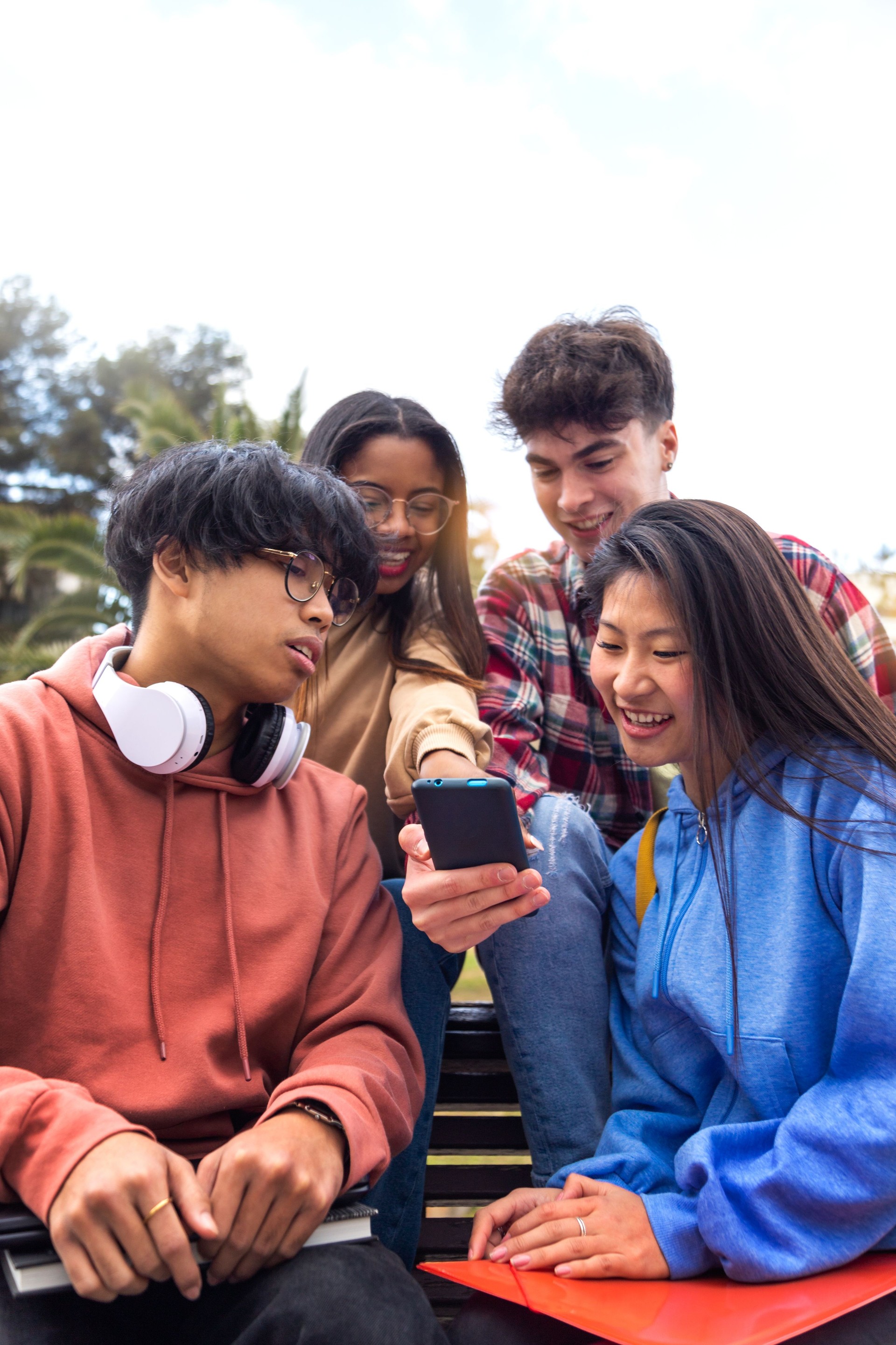 Multiracial happy young college student friends look at mobile phone. Teenagers using smartphone outdoor. Vertical.