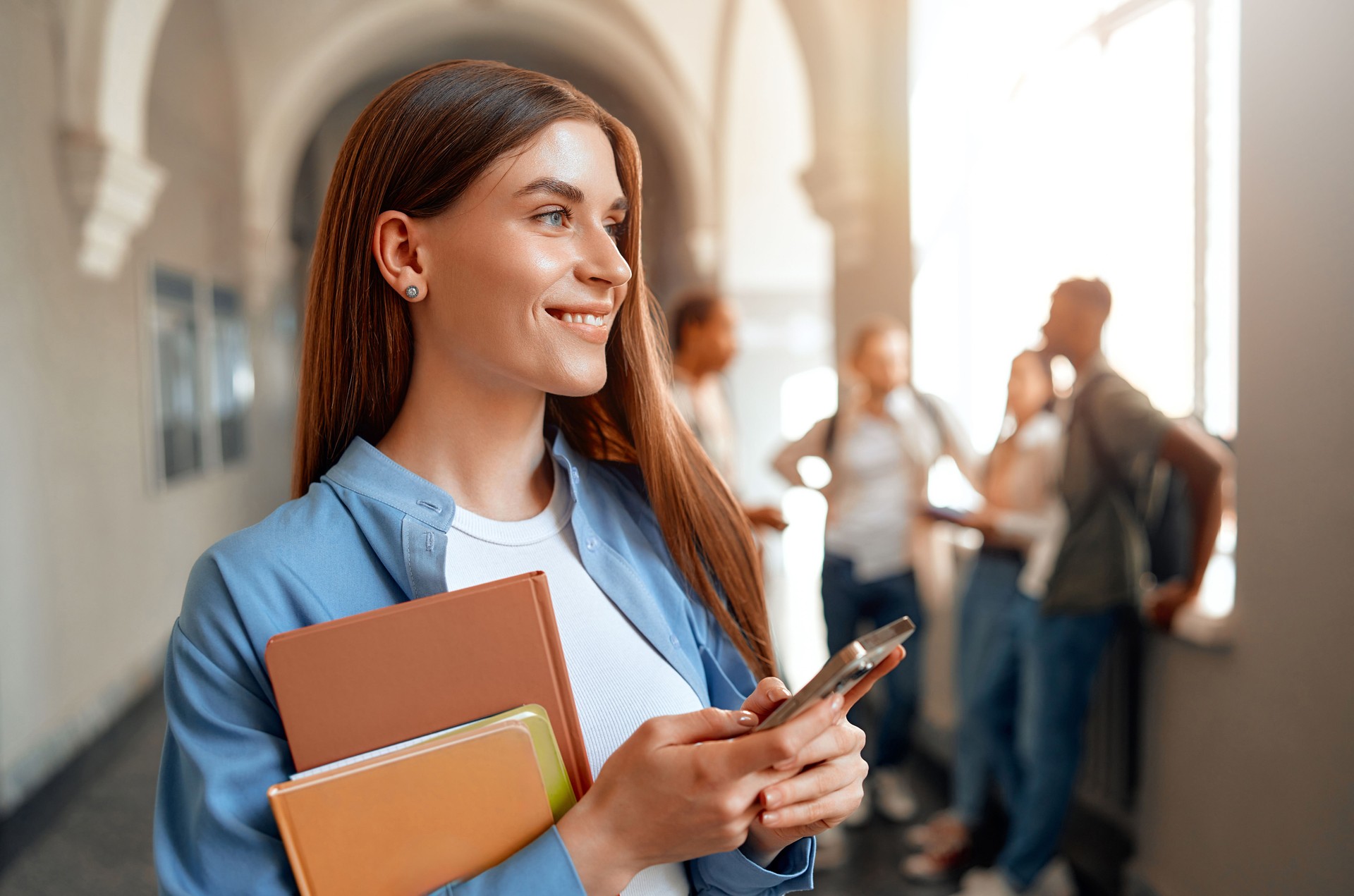 A student texts on a smartphone in a university hallway with classmates in the background