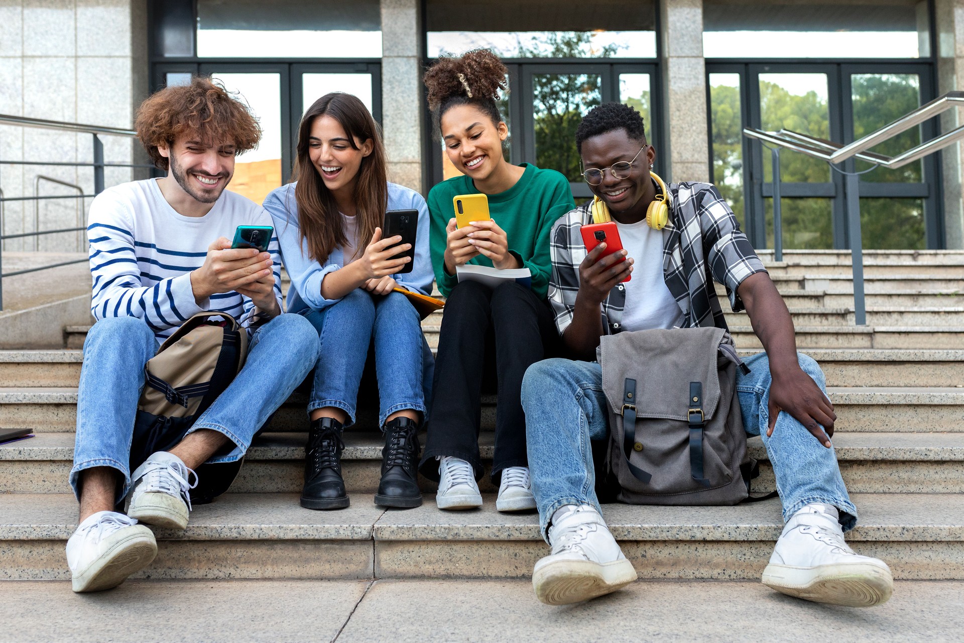 Multiracial college student friends look mobile phone laughing together. University students using smartphone outdoors.